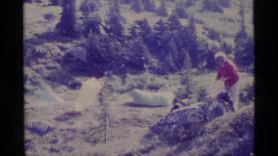1977 Alaska home-movie frame with a woman in a red jacket handling campsite gear near white tents and low alpine vegetation.