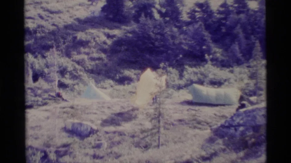 1977 Alaska frame showing white tents, windblown fabric, rocks, and trees across an exposed mountain campsite.