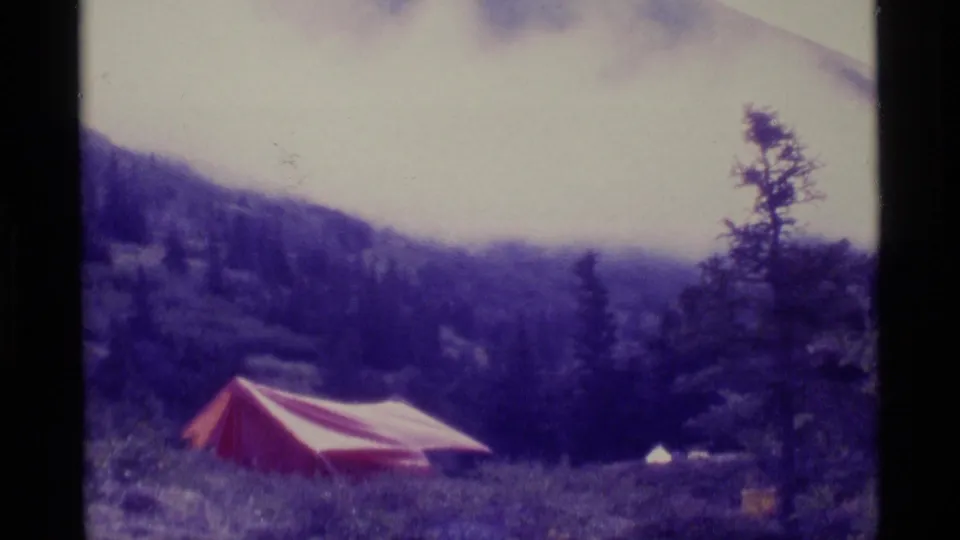 1977 Alaska 8mm frame with a red and white tent below a misty mountain and evergreen trees.