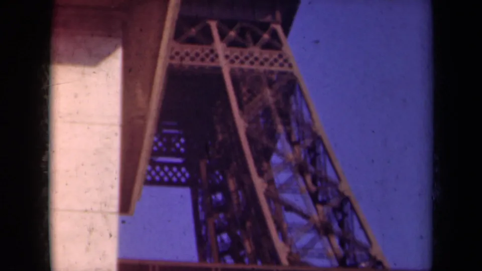 1939 Paris home-movie view looking up at Eiffel Tower iron beams near a pale overhang, with saturated blue sky.