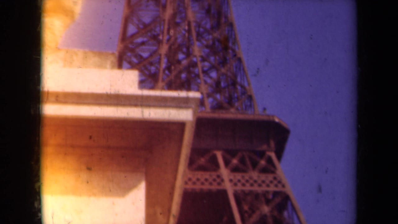 1939 Paris home-movie frame showing the Eiffel Tower lattice rising near a stone statue under a blue-purple sky.
