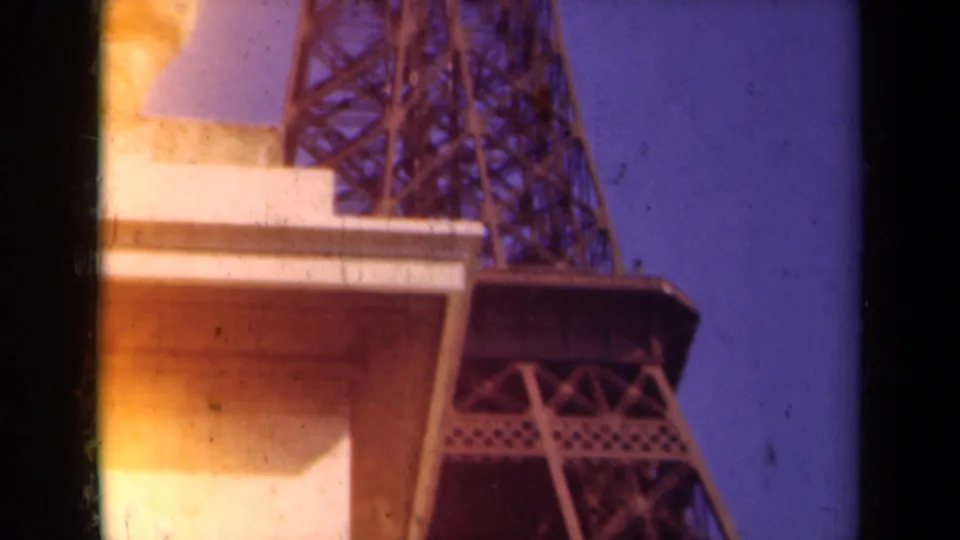 1939 Paris home-movie frame showing the Eiffel Tower lattice rising near a stone statue under a blue-purple sky.