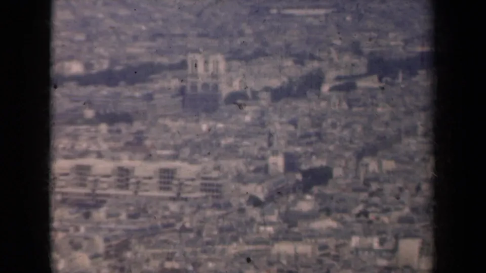 1939 Paris cityscape from above, with twin cathedral-like towers, dense buildings, and muted color from 8mm film.