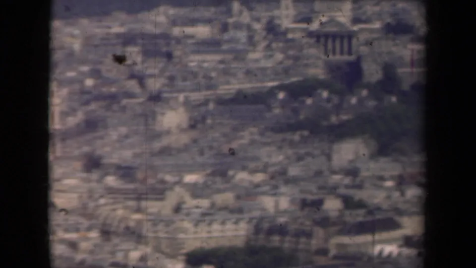 Soft 8mm frame of Paris from a high viewpoint, showing rooftops, dark green park areas, and distant monumental forms.