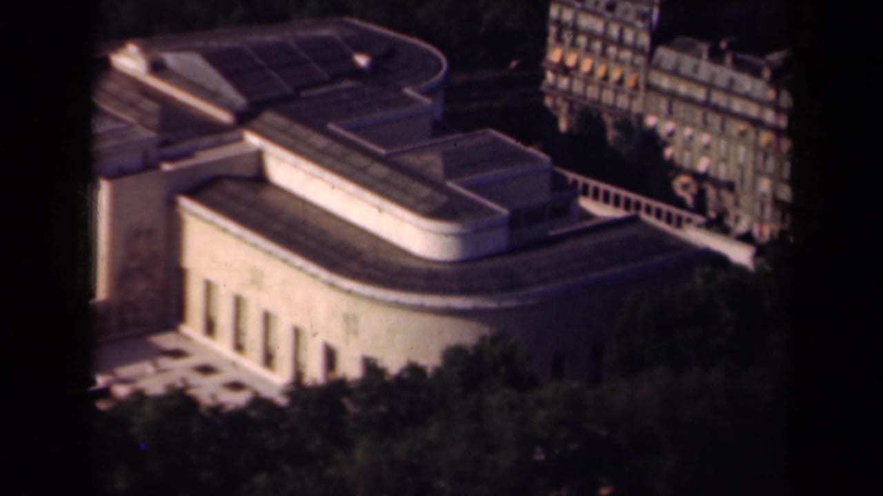 Aerial view of a large curved building with flat roofs, columns, trees, and adjacent city blocks.