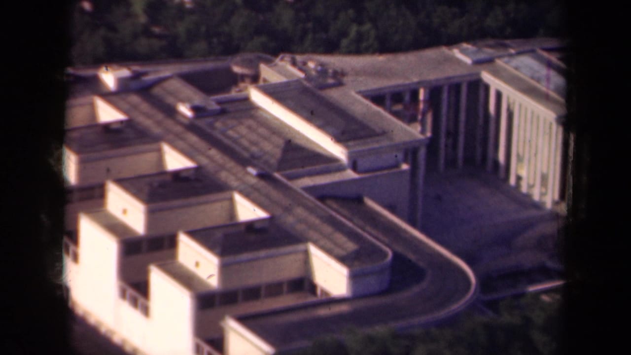 1939 Paris aerial frame with a modernist building complex, rooftop sections, pale facades, and dark trees beyond.