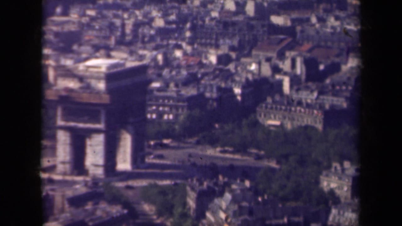 Soft 8mm aerial frame of the Arc de Triomphe in Paris, with traffic below and compact urban buildings in the background.