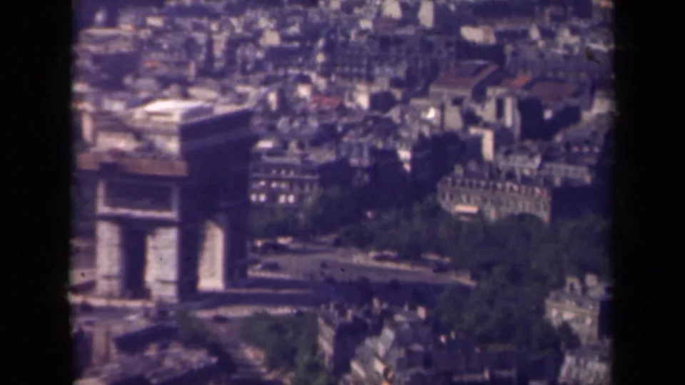 Soft 8mm aerial frame of the Arc de Triomphe in Paris, with traffic below and compact urban buildings in the background.