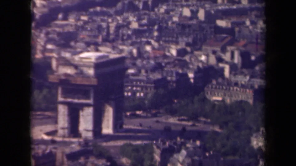 Aerial home-movie frame of the Arc de Triomphe in Paris, with moving cars below and the surrounding city softened by vintage film grain.