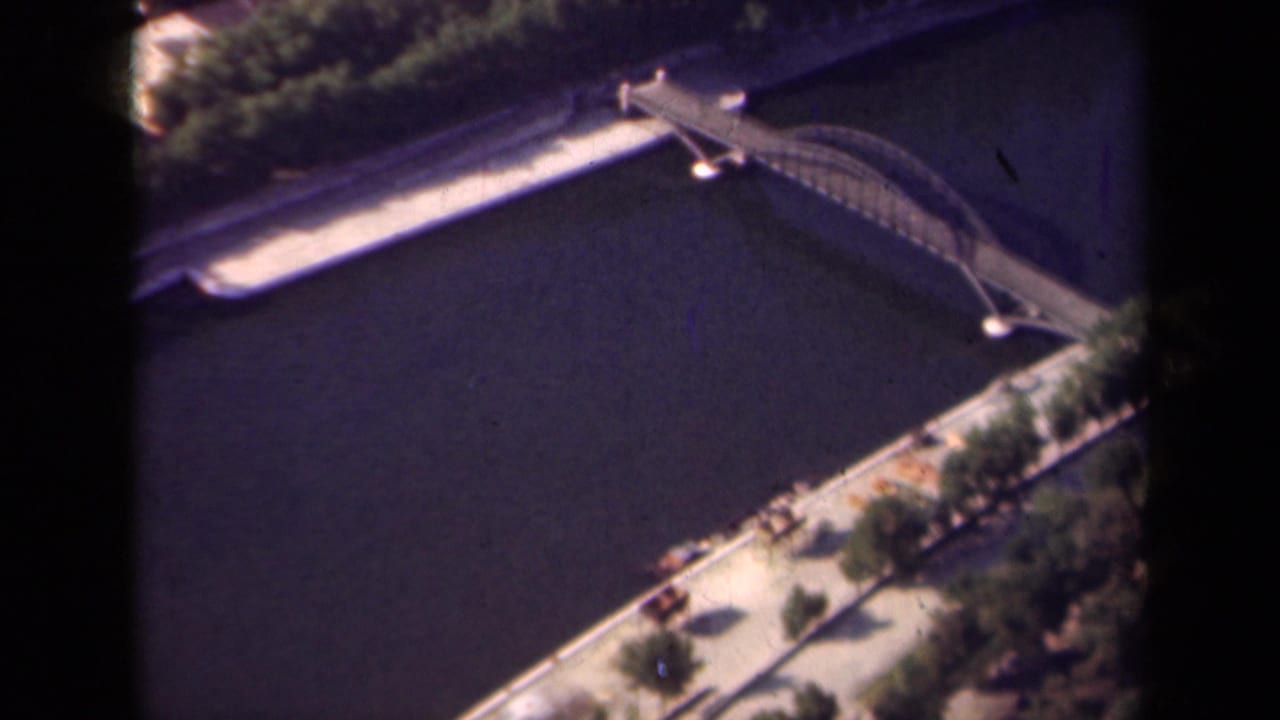 1939 Paris home-movie frame showing an elevated view of an arched bridge over a broad river, bordered by trees and embankment paths.