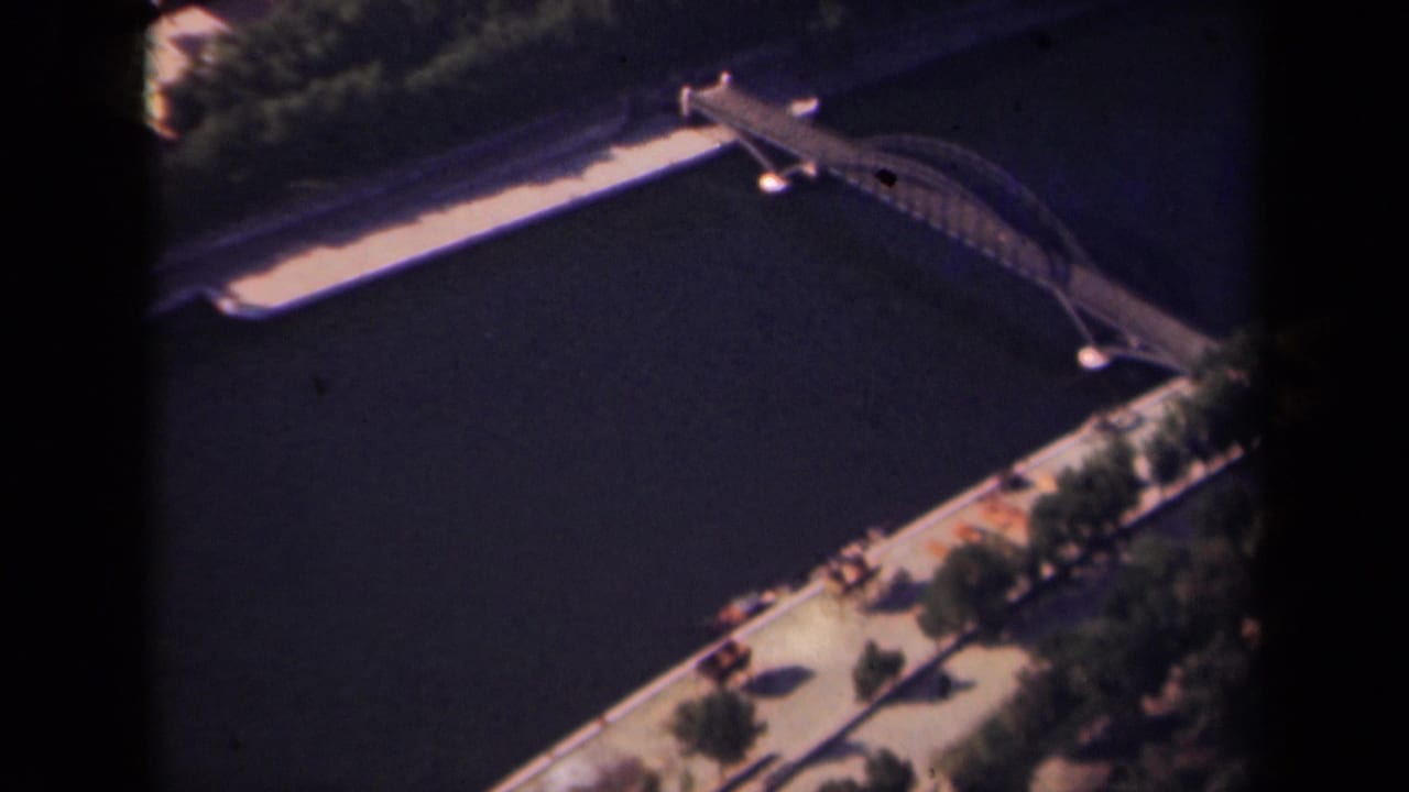 Aerial 8mm view in Paris, 1939, with an arched bridge, dark river surface, trees, and a light-toned riverside walkway.