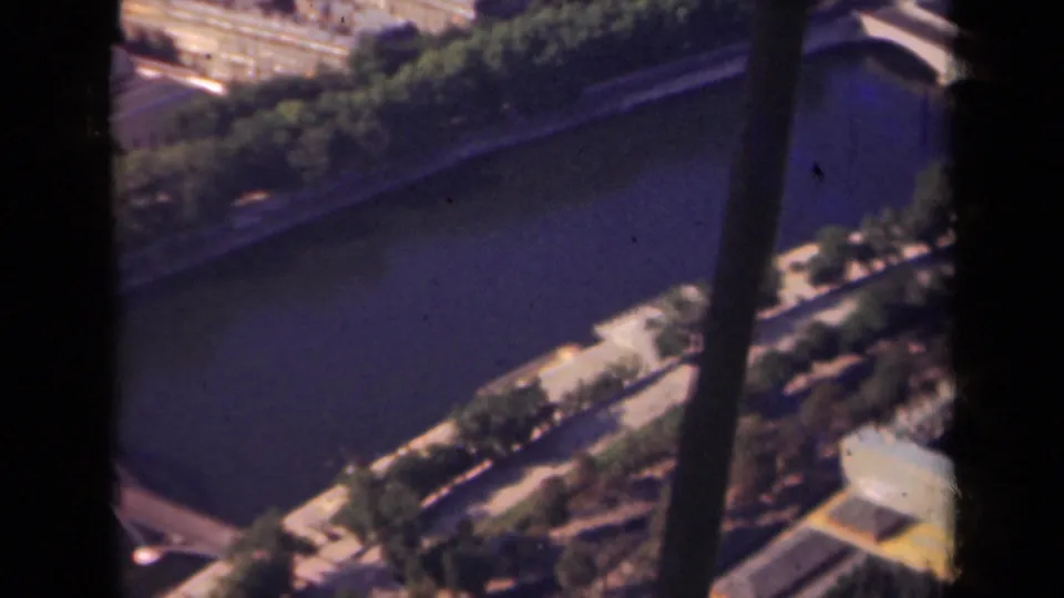 Paris riverfront seen from above, with the Seine, a bridge crossing, quayside trees, and dense urban blocks in soft 8mm color.