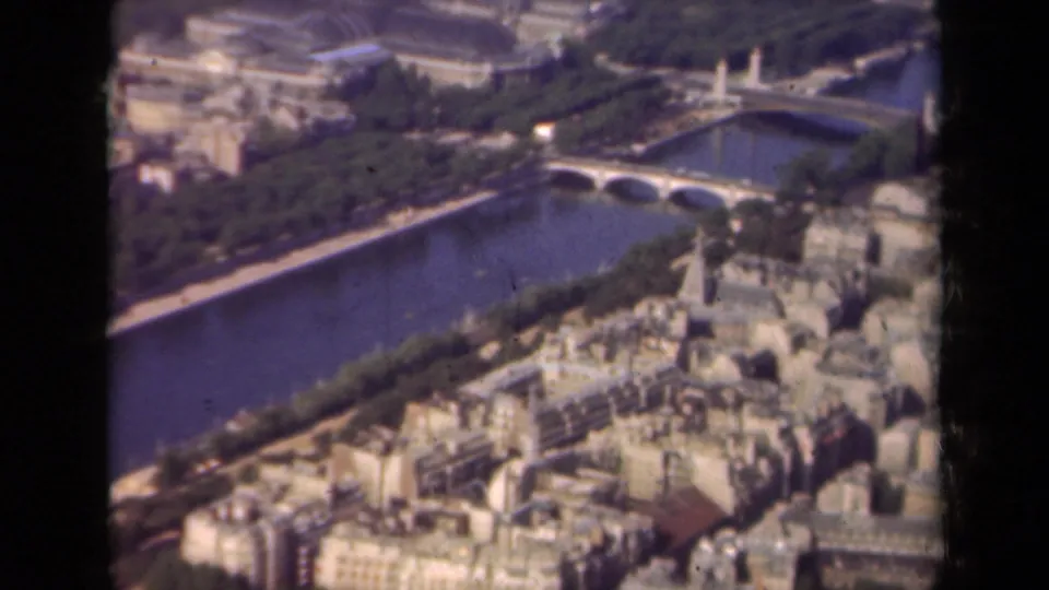 1939 Paris aerial frame with a multi-arch Seine bridge, embankments, dense buildings, and soft 8mm color grain.
