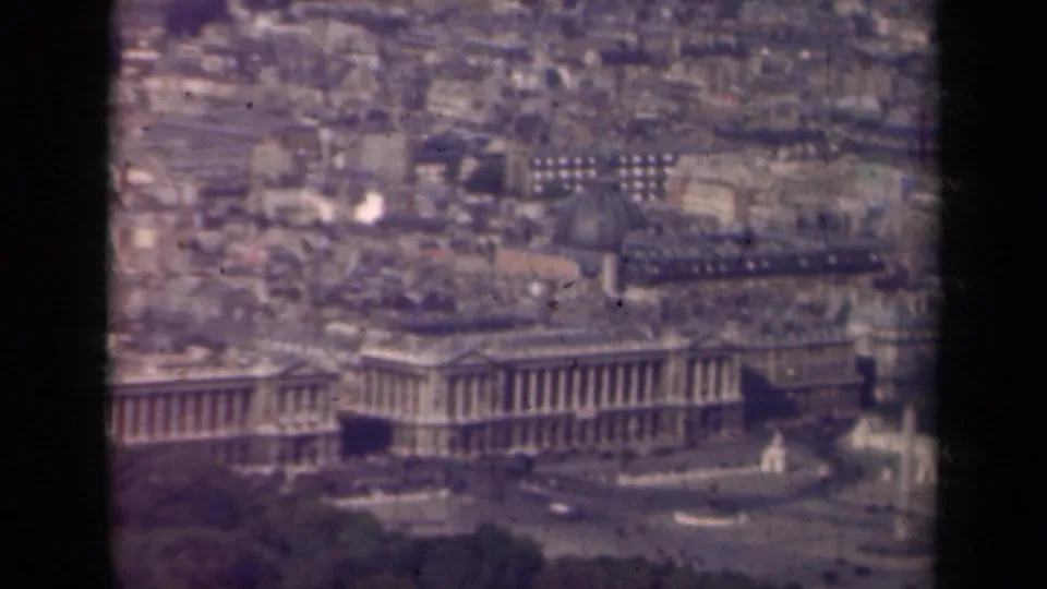 1939 Paris aerial frame showing a long colonnaded building below dense rooftops, with trees, roads, and fountains nearby.