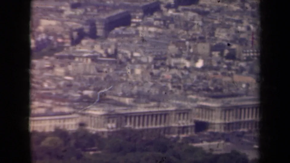 1939 Paris aerial frame with rooftops, a large colonnaded building, tree cover, roads, and pale fountains in soft focus.