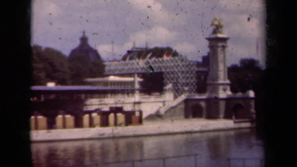 1939 Paris home-movie frame showing water, a stone bridge or quay, a domed building, glass-roofed structure, trees, and a gilded statue column.