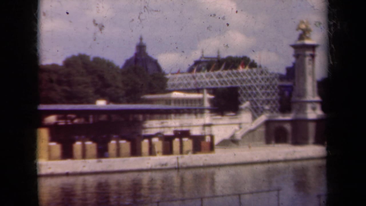 Waterfront architecture with bridge steps, glass-roofed structure, domed roof, trees, and statue-topped column.