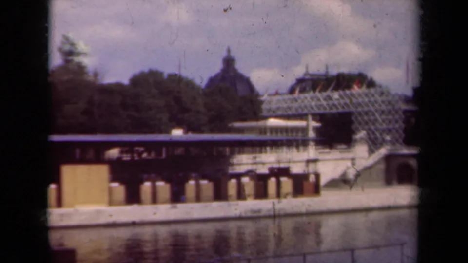 1939 Paris home-movie frame during a pan, with foreground water, railings, trees, glass-roofed architecture, and a domed roof beyond.