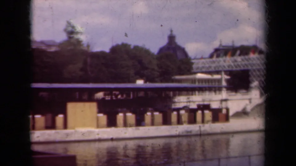 1939 Paris frame with soft focus and film grain, showing water, a bridge or quay, trees, a glass-roofed building, and a domed roof in the background.