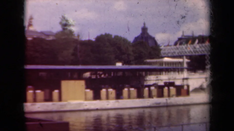 1939 Paris home-movie frame showing a riverfront structure, leafy bank, and distant dome beyond the trees.