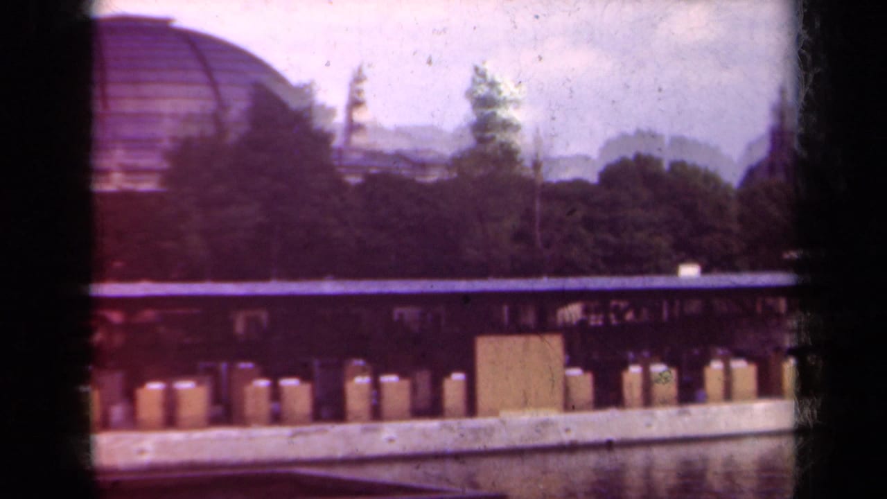 Stone quay and bollards along water with trees and a domed roof beyond.