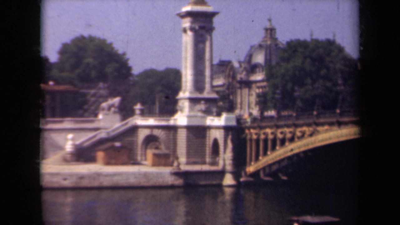 Ornate arched bridge over a river with stone pylons, trees, and buildings beyond.