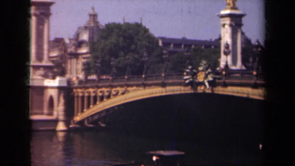 1939 Paris frame showing the ornate bridge and river from a visitor viewpoint, with small distant figures near the railing and trees behind.