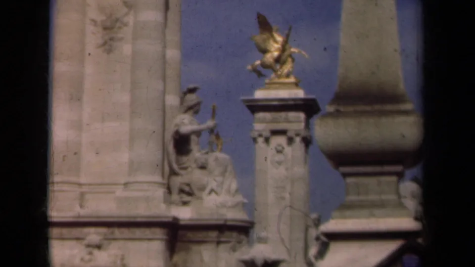 1939 Paris frame focusing on Pont Alexandre III ornament, with a seated sculpture, gilded winged figure, pale stonework, and blue sky.