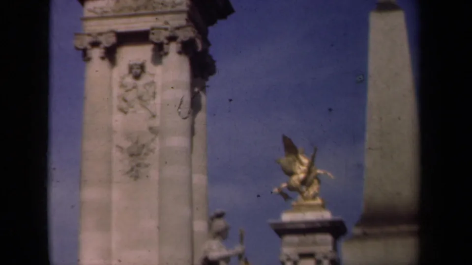 1939 Paris home-movie frame showing a carved stone monument column, relief ornament and a distant gilded winged horse sculpture against blue sky.