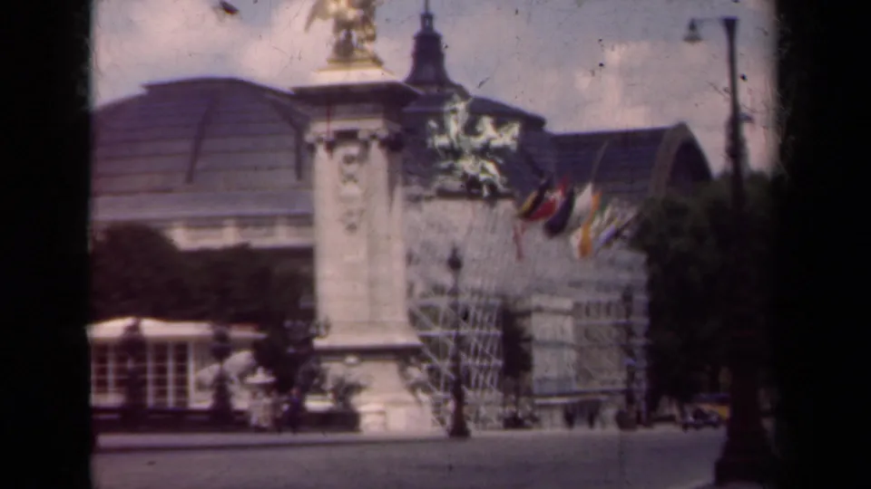 1939 Paris 8mm frame showing the statue on a tall pedestal near scaffolding, flags, and a broad roofed building.