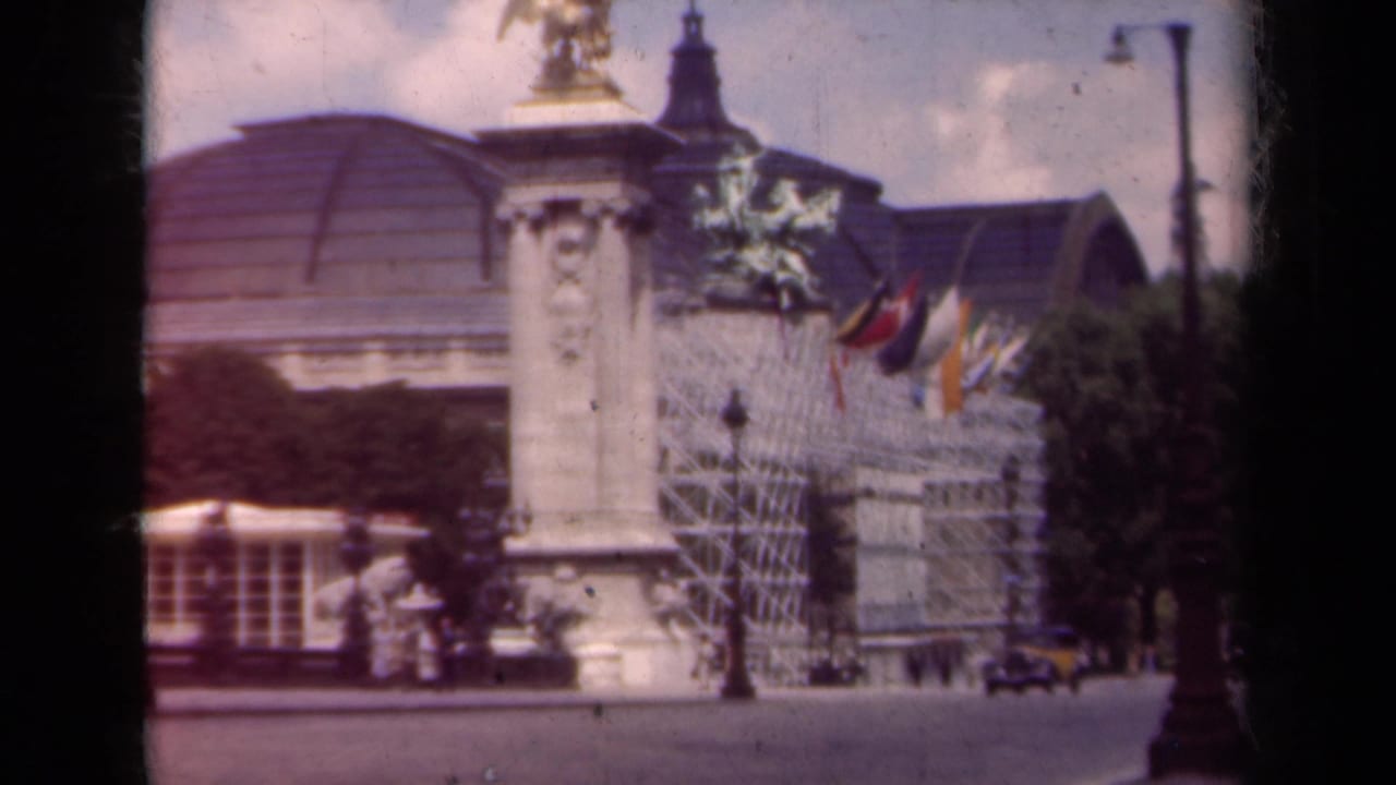 Ornate Paris street building with flags, statue column, cars, pedestrians, and streetlamps.
