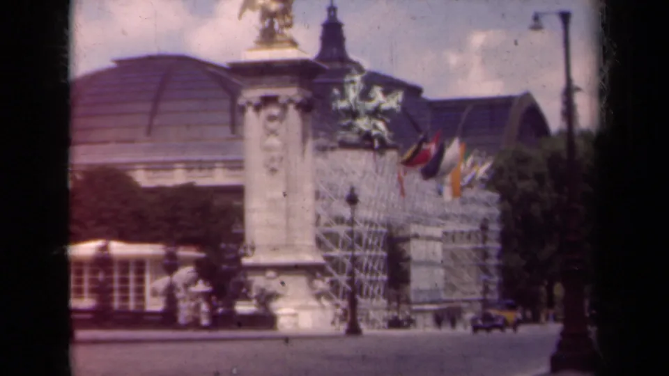 1939 Paris street view showing an ornate facade with international flags, a sculptural column, cars, pedestrians, and lamps.