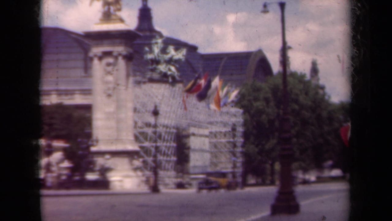 1939 Paris boulevard scene with a flag-lined ornate building, statue column, passing cars, streetlamp, and trees.
