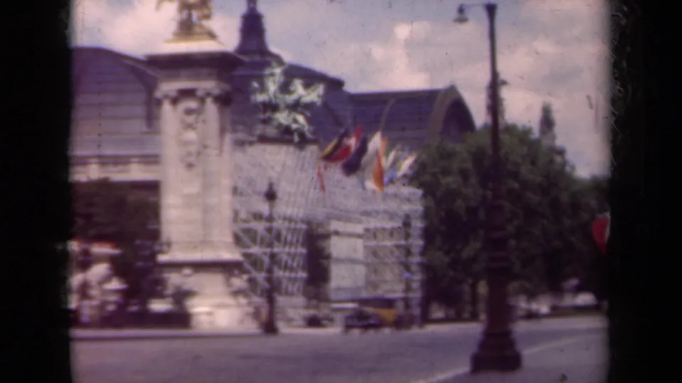 1939 Paris boulevard scene with a flag-lined ornate building, statue column, passing cars, streetlamp, and trees.