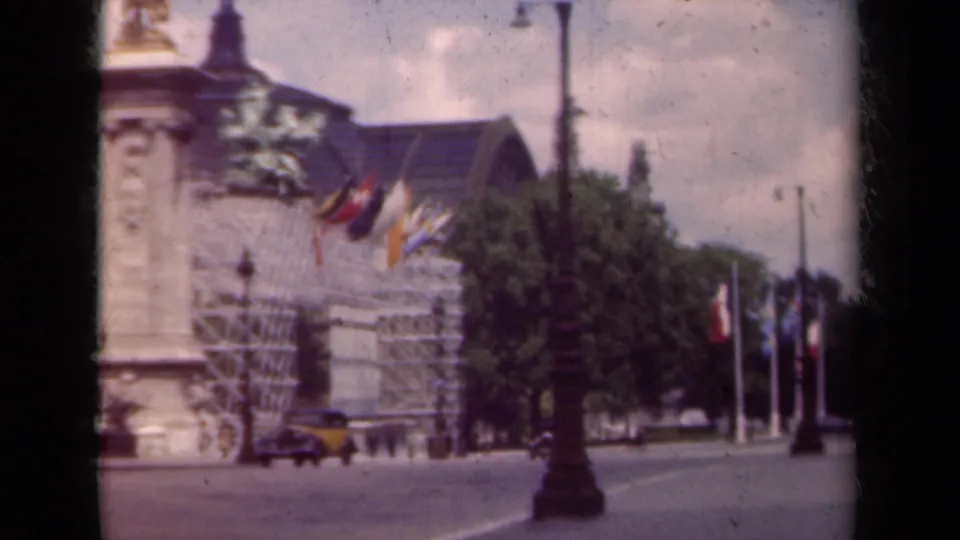 1939 Paris home-movie frame with flags on an ornate building, a streetlamp, trees, pedestrians, and a yellow car.
