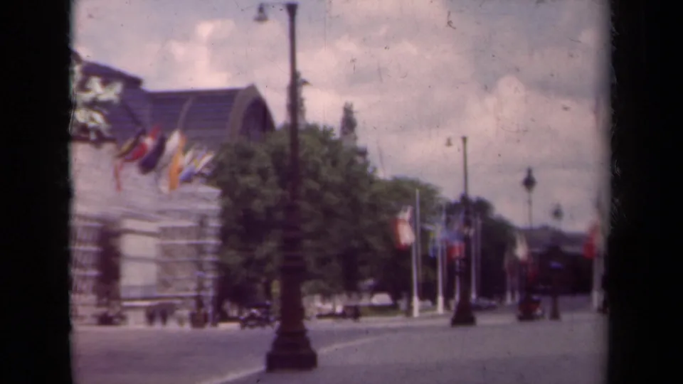1939 Paris boulevard view with a decorated facade, rows of flags, trees, streetlamps, cars, and distant pedestrians.