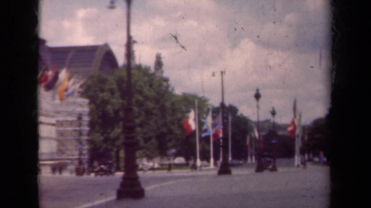 Flag-lined Paris boulevard with trees, lampposts, a large building, and a small automobile.