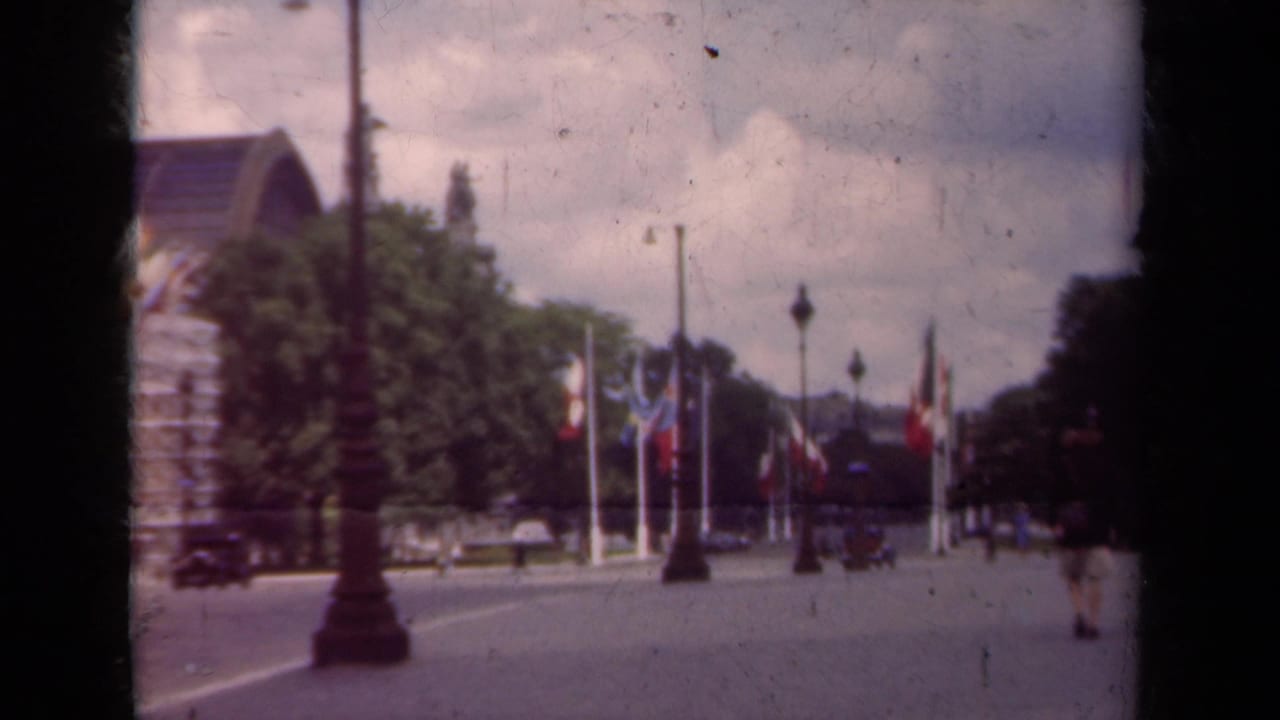 1939 Paris boulevard seen from a moving home-movie camera, with flagpoles, trees, lampposts, pedestrians, and distant vehicles.