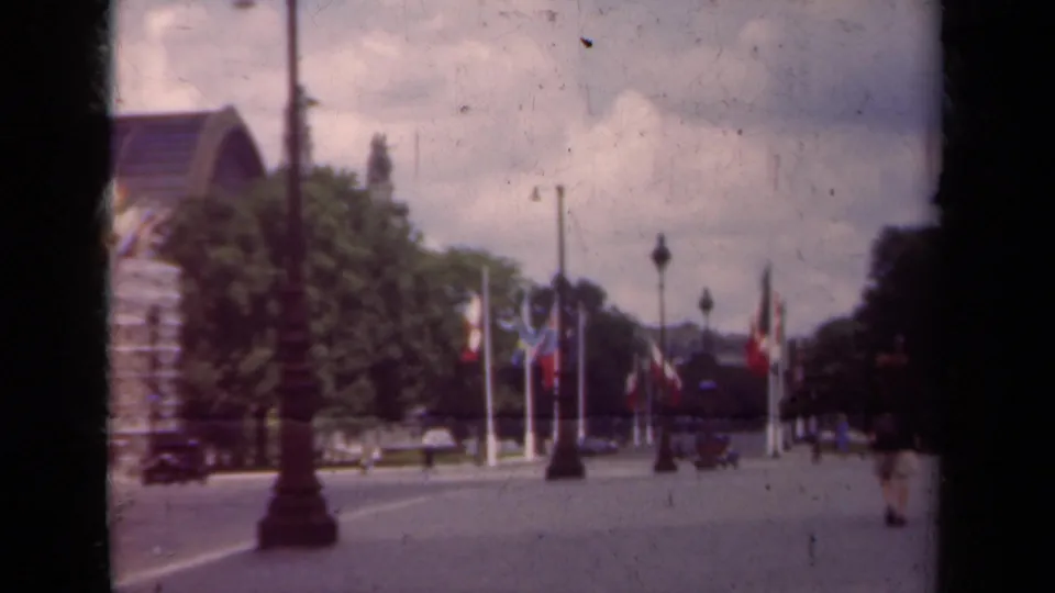 1939 Paris boulevard seen from a moving home-movie camera, with flagpoles, trees, lampposts, pedestrians, and distant vehicles.