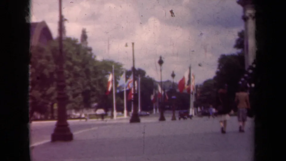 1939 Paris street scene with multiple flags, tall lampposts, trees, pedestrians, and a partially visible large building at frame edge.