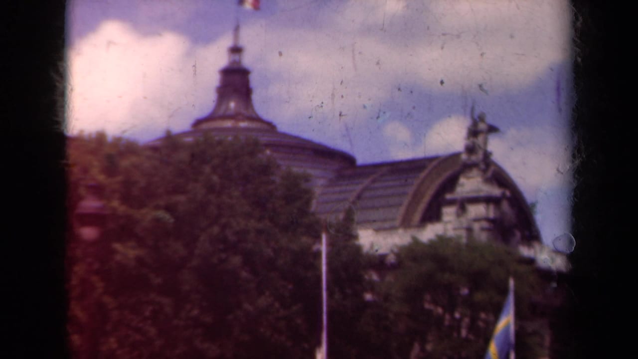 1939 Paris home-movie frame showing a grand stone building roofline behind trees, with flagpoles and soft color 8mm texture.