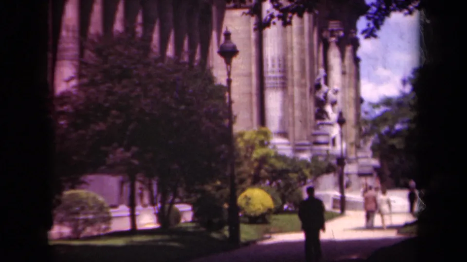 Late-1930s Paris home-movie frame showing people near a monumental stone facade, with statue, trees, shrubs, and flag colors at the edge.