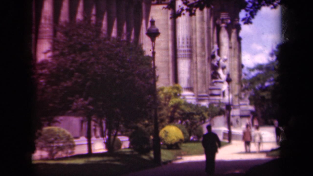 1939 Paris home-movie frame showing a suited man on a sunlit path near a columned stone building, statue, trees, shrubs, and lamppost.
