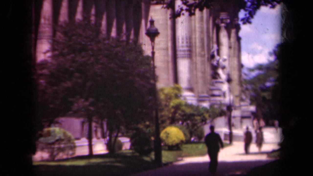 1939 Paris footage frame with a dark-suited figure walking toward a monumental facade, with other pedestrians near the entrance path.