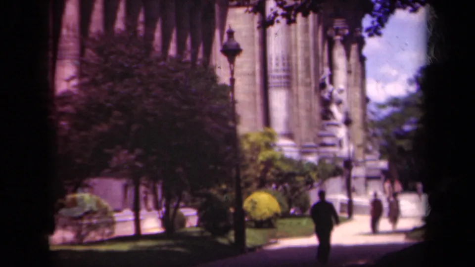 1939 Paris footage frame with a dark-suited figure walking toward a monumental facade, with other pedestrians near the entrance path.