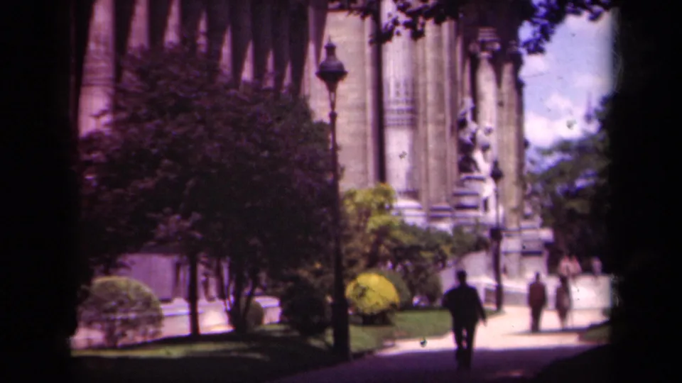1939 Paris home-movie frame showing a man walking through partial shade toward a columned building with statue and greenery.