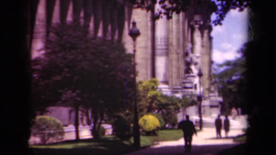 Final 1939 Paris frame showing a suited pedestrian and distant figures near a monumental columned building, statue, lamppost, and greenery.