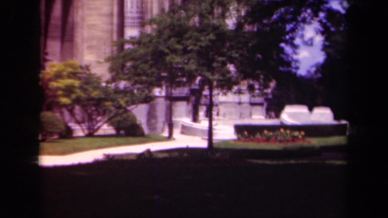 Stone building partly seen behind trees, lawn, walkway, and a low flower bed.