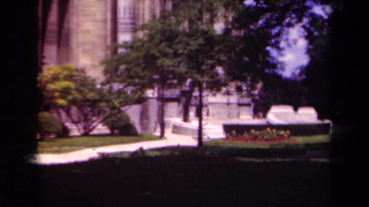 Paris 1939 home-movie frame showing a formal stone facade beyond trees, lawn, path, and a low flower bed.