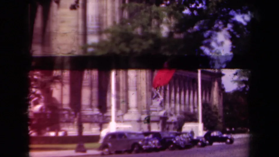 1939 Paris street view with a columned official-looking building, flagpoles, flags, and several parked classic cars.